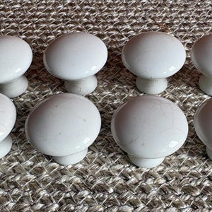 May include: A set of eight white ceramic cabinet knobs arranged in two rows of four. Each knob features a smooth, rounded top with a subtle crackle glaze finish and a tapered base, providing a uniform appearance for furniture hardware applications.
