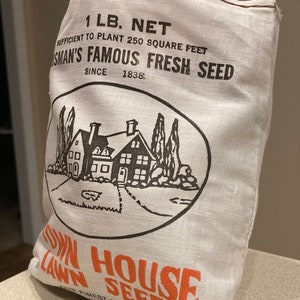 May include: A white burlap bag with brown stitching, labeled "1 LB. NET" and "SUFFICIENT TO PLANT 250 SQUARE FEET",  "OSMAN'S FAMOUS FRESH SEED", "SINCE 1838". The bag features a black and white illustration of a house with trees and a lawn. The bottom of the bag is labeled "TOWN HOUSE LAWN SEED" in orange.