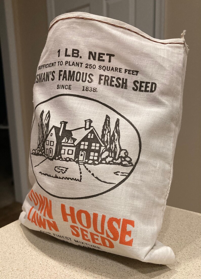 May include: A white burlap bag with brown stitching, labeled "1 LB. NET" and "SUFFICIENT TO PLANT 250 SQUARE FEET",  "OSMAN'S FAMOUS FRESH SEED", "SINCE 1838". The bag features a black and white illustration of a house with trees and a lawn. The bottom of the bag is labeled "TOWN HOUSE LAWN SEED" in orange.