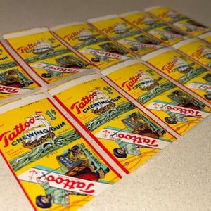 May include: A collection of vintage Tattoo Chewing Gum wrappers. The wrappers are yellow with red and blue text and illustrations of a ship, sailor, and anchor. The text includes "Tattoo Chewing Gum" and "1&cent;".