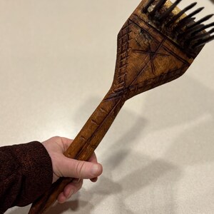 Puede incluir: Un peine de madera tallado a mano con un dise&ntilde;o de estrella y geom&eacute;trico. El peine presenta cerdas oscuras y un mango largo. La madera tiene un tono marr&oacute;n c&aacute;lido y el peine se sostiene en una mano.