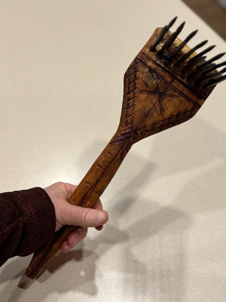 Puede incluir: Un peine de madera tallado a mano con un dise&ntilde;o de estrella y geom&eacute;trico. El peine presenta cerdas oscuras y un mango largo. La madera tiene un tono marr&oacute;n c&aacute;lido y el peine se sostiene en una mano.