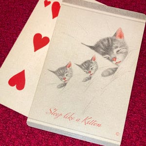 Puede incluir: Una carta de juego con el siete de corazones junto a una carta con un dibujo de tres gatitos durmiendo. Los gatitos son grises con orejas y narices rosas. El texto "Sleep like a Kitten" est&aacute; escrito en cursiva en la tarjeta.