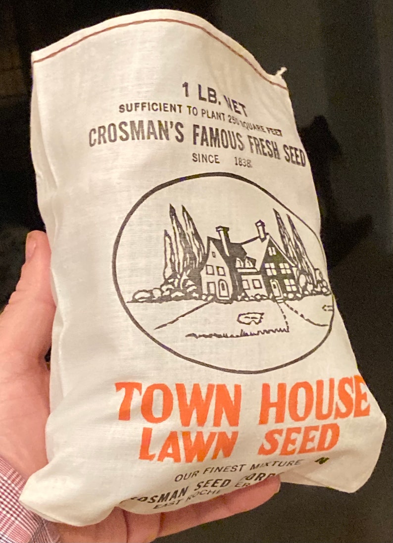 May include: A white burlap bag with black and orange text that reads "Crosman's Famous Fresh Seed" and "Town House Lawn Seed". The bag is 1 lb. net and is sufficient to plant 250 square feet. The bag also features a black and white illustration of a house with trees and a driveway.