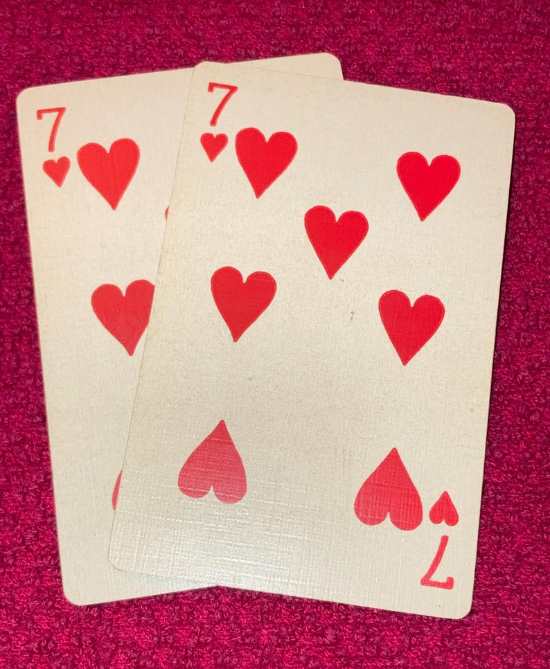 Puede incluir: Se muestran dos cartas de juego, ambas el siete de corazones. Las cartas tienen un fondo blanco con s&iacute;mbolos de coraz&oacute;n rojos y el n&uacute;mero siete en las esquinas. Las cartas parecen ligeramente desgastadas.