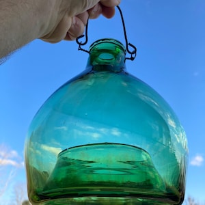 May include: A translucent emerald green glass bottle with a wide, rounded body and a narrow neck. The bottle features a thick, indented base and a dark metal wire handle attached to the rim, allowing it to be suspended or carried by hand.