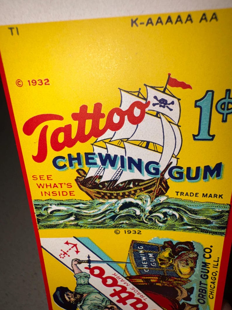May include: Vintage "Tattoo Chewing Gum" packaging. The yellow background features a pirate ship illustration with a skull and crossbones flag, the text "Tattoo Chewing Gum", and the price "1&cent;". Includes the text "See What's Inside" and "Trade Mark".