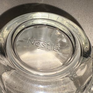 Może przedstawiać: Przezroczysty szklany kubek z uchwytem, z wytłoczonym słowem "Nestle" na dnie. Kubek jest przezroczysty, przepuszcza światło i wydaje się czysty. Kubek jest prawdopodobnie przeznaczony do napoj&oacute;w.