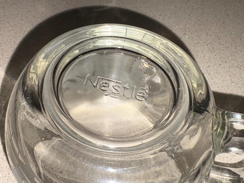 Może przedstawiać: Przezroczysty szklany kubek z uchwytem, z wytłoczonym słowem "Nestle" na dnie. Kubek jest przezroczysty, przepuszcza światło i wydaje się czysty. Kubek jest prawdopodobnie przeznaczony do napoj&oacute;w.