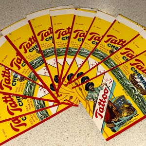 May include: A fan of vintage Tattoo Chewing Gum wrappers. The wrappers are yellow with red and white text and graphics. The wrappers feature a sailor and a ship. The wrappers are from 1932.