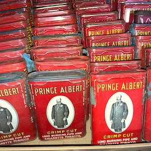 May include: A collection of vintage Prince Albert tobacco tins. The tins are red with gold lettering and feature a man in a suit and top hat. The text on the tins reads "Prince Albert Crimp Cut Long Burning Pipe and Cigarette Tobacco".
