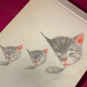 Puede incluir: Primer plano de una tela clara con un patr&oacute;n repetido de tres gatitos grises con orejas y narices rojas. Los gatitos est&aacute;n representados con los ojos cerrados, dando una apariencia pac&iacute;fica.