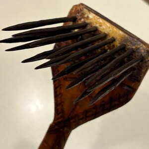 Puede incluir: Un peine de madera con p&uacute;as oscuras y puntiagudas. El peine tiene un mango y una cabeza cuadrada con m&uacute;ltiples p&uacute;as. La madera tiene un color marr&oacute;n oscuro con un tono amarillento m&aacute;s claro en la cabeza.