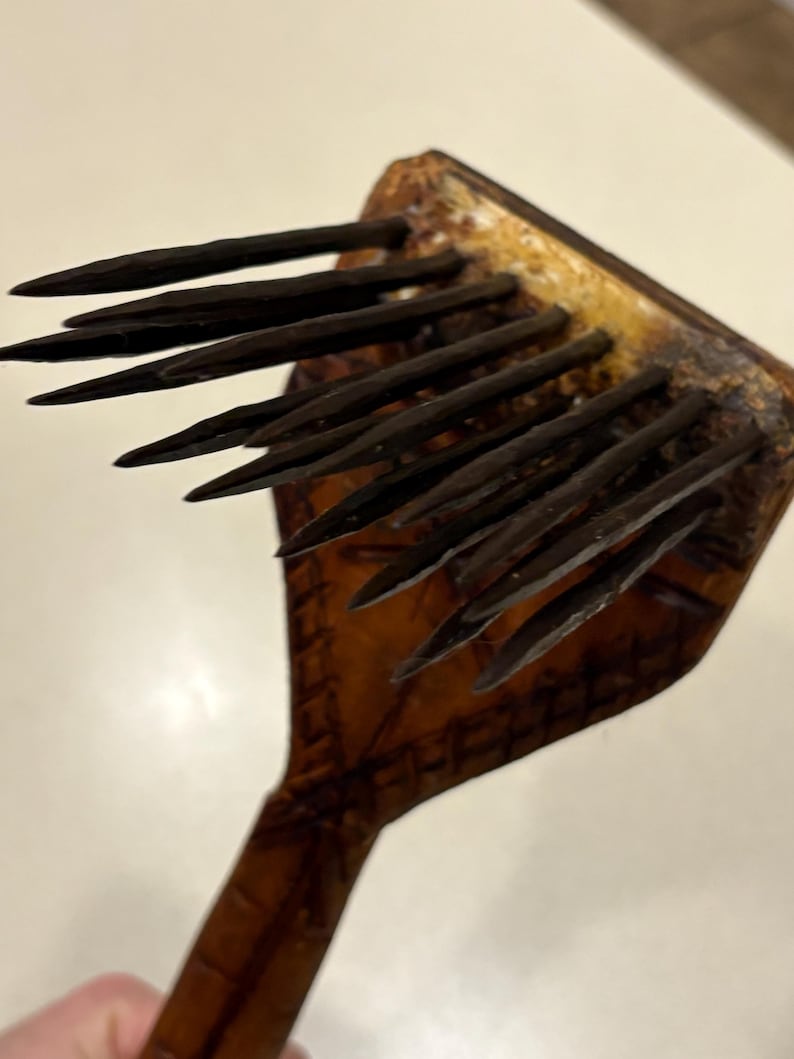 Puede incluir: Un peine de madera con p&uacute;as oscuras y puntiagudas. El peine tiene un mango y una cabeza cuadrada con m&uacute;ltiples p&uacute;as. La madera tiene un color marr&oacute;n oscuro con un tono amarillento m&aacute;s claro en la cabeza.