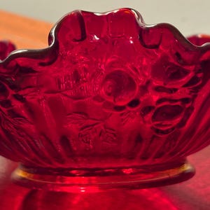 May include: A close-up of a ruby red glass bowl with a ruffled edge. The bowl features a floral pattern and a gold-colored base. The glass is translucent, allowing light to pass through, highlighting the intricate details of the design.