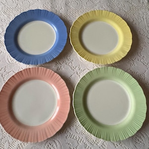 May include: Four colorful ceramic plates. Each plate has a white center and a scalloped rim in a different pastel color: blue, yellow, pink, and green. The plates are arranged on a white lace tablecloth.