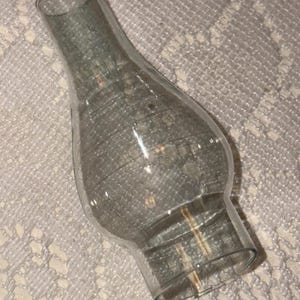 May include: A clear glass lamp chimney with a flared base and a rounded body. The chimney is slightly curved and tapers towards the top, with a smooth, open rim. The glass has a slight greenish tint.