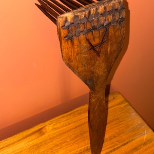 Puede incluir: Un peine de pelo de madera vintage con mango y p&uacute;as largas y finas. La cabeza del peine est&aacute; adornada con tachuelas met&aacute;licas y un s&iacute;mbolo de estrella tallado. La madera tiene un aspecto c&aacute;lido y envejecido, lo que sugiere que es una antig&uuml;edad.