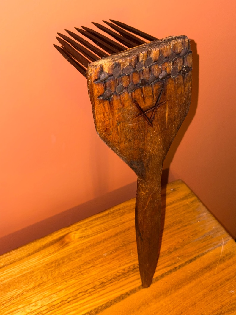 Puede incluir: Un peine de pelo de madera vintage con mango y p&uacute;as largas y finas. La cabeza del peine est&aacute; adornada con tachuelas met&aacute;licas y un s&iacute;mbolo de estrella tallado. La madera tiene un aspecto c&aacute;lido y envejecido, lo que sugiere que es una antig&uuml;edad.
