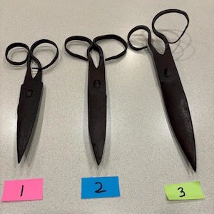 May include: Three dark brown, hand-forged iron scissors of varying sizes arranged in a row. Each pair features a singular loop handle and a pointed blade. Below each pair of scissors is a small rectangular paper label numbered 1, 2, and 3.