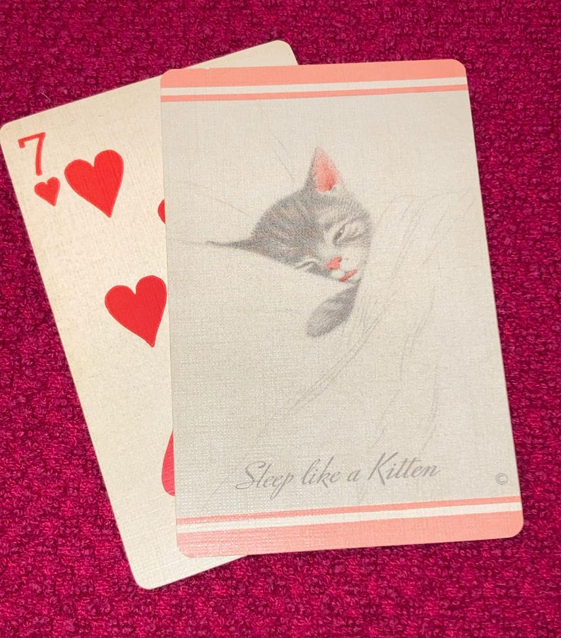 Puede incluir: Dos cartas de juego sobre un fondo rosa. Una carta muestra el n&uacute;mero siete y tres corazones rojos. La otra carta presenta un gatito gris durmiendo con el texto "Sleep like a Kitten" en un borde rosa claro.