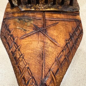 Puede incluir: Primer plano de un peine de madera con un dise&ntilde;o tallado. El peine es de color marr&oacute;n oscuro con un patr&oacute;n en forma de cruz y detalles tejidos. Los dientes del peine son oscuros y se extienden hacia arriba.