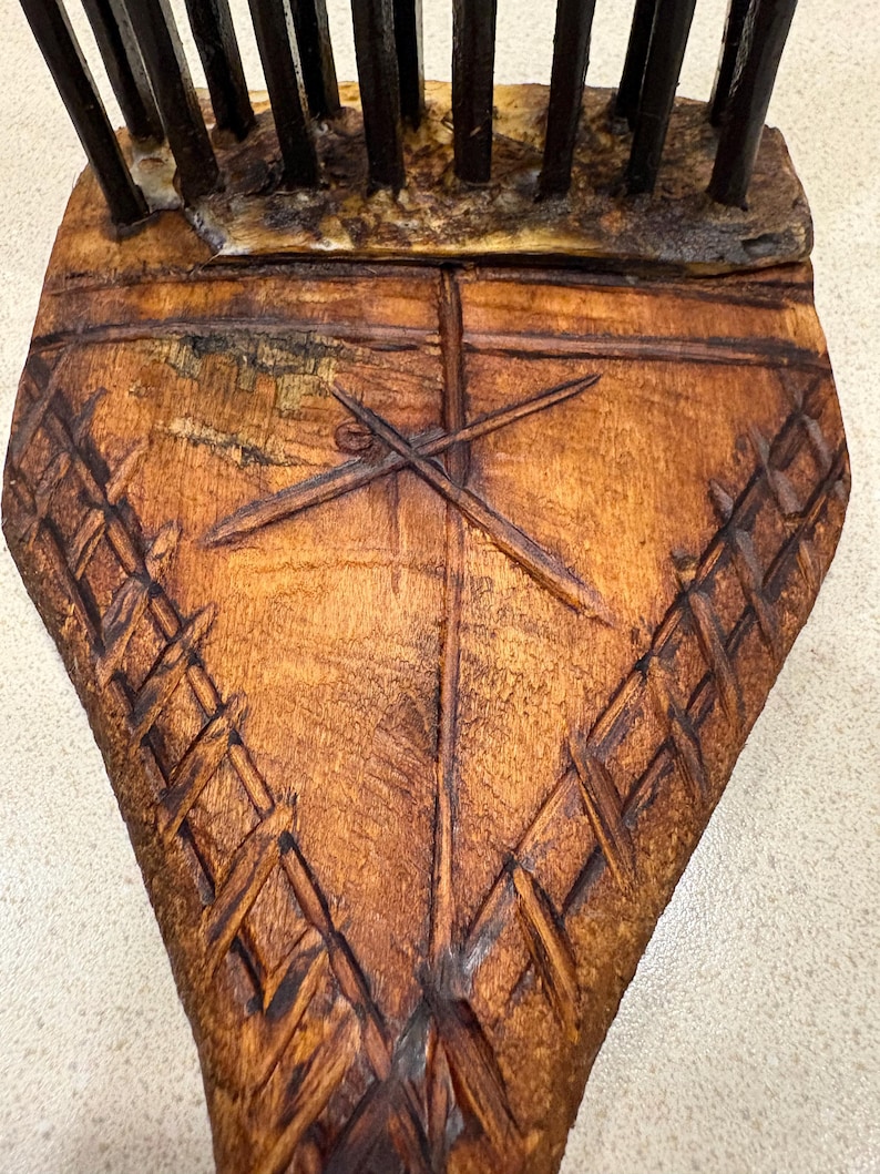 Puede incluir: Primer plano de un peine de madera con un dise&ntilde;o tallado. El peine es de color marr&oacute;n oscuro con un patr&oacute;n en forma de cruz y detalles tejidos. Los dientes del peine son oscuros y se extienden hacia arriba.