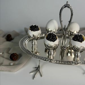 Könnte beinhalten: Ein silbernes Serviertablett mit Eierschalenbechern, die mit schwarzem Kaviar gefüllt sind. Das Tablett hat einen dekorativen Rand und einen Griff. Die Eierschalenbecher stehen auf verzierten Silberständern. Der Hintergrund ist ein neutrales Grau.