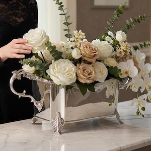 May include: A silver rectangular flower vase filled with white and tan roses, orchids, and greenery. The vase has ornate handles and feet. The arrangement sits on a marble-topped table.