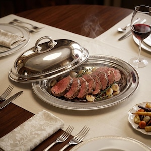 Puede incluir: Una fuente de plata con tapa abovedada, que contiene carne asada en rodajas y cebollas asadas. La fuente está sobre un mantel blanco, con una copa de vino tinto y cubiertos. También hay un plato de verduras asadas.