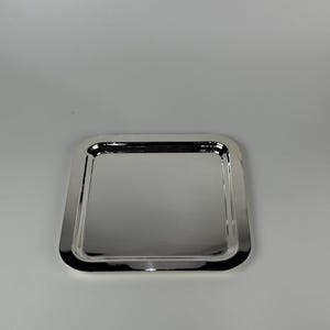 May include: A polished, square, silver tray with rounded corners. The tray has a reflective surface and a slightly raised edge. The tray is empty and sits on a white surface.