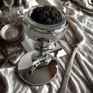 Puede incluir: Un juego de servir caviar plateado con un cuenco de cristal lleno de caviar oscuro. El juego incluye una tapa con un asa en forma de pez, una cuchara de servir y una base plateada. El juego está sobre un mantel blanco con cubertería.