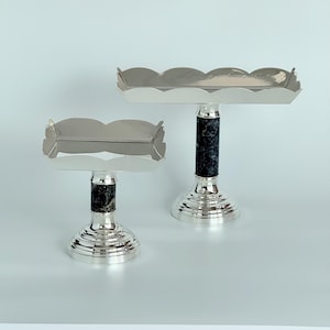 Könnte beinhalten: Zwei silberfarbene Serviertabletts mit gewellten Kanten. Jedes Tablett hat einen dunklen, marmorierten Säulensockel und einen abgestuften, kreisförmigen Fuß. Die Tabletts werden vor einem hellblauen Hintergrund präsentiert.