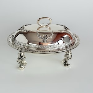 May include: A silver serving dish with a domed lid and floral embellishments. The dish has a decorative rim and is supported by four ornate feet. The lid features a loop handle. The dish is likely made of silver plate.