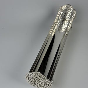 Pode incluir: Um objeto prateado e alongado com um design decorativo vazado em ambas as extremidades. O corpo tem ranhuras verticais, e a parte superior e inferior apresentam um padrão detalhado em forma de renda. O objeto parece ser de metal.