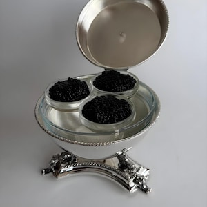 May include: A silver caviar server with three glass dishes filled with black caviar. The server has a decorative base and a lid that is partially open. The server is on a white surface.