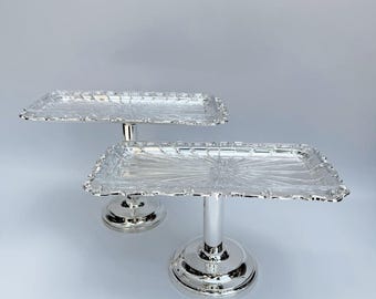 Puede incluir: Dos soportes rectangulares plateados para pasteles. Los soportes tienen un borde decorativo festoneado y un patrón de estrella en la superficie. Cada soporte está sostenido por una base redonda y un tallo cilíndrico. Ideales para postres.