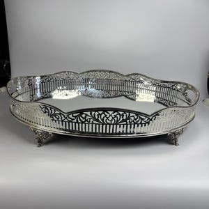 Puede incluir: Una bandeja de servicio ovalada de color plateado con una base de espejo y un borde decorativo calado. La bandeja presenta un intrincado patrón de filigrana en los laterales, bordes festoneados y cuatro patas ornamentadas.