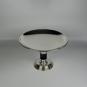 Op de afbeelding: Een zilverkleurige voetstandaard met een brede, cirkelvormige, reflecterende bovenplaat. De centrale steunkolom bevat een donker, getextureerd cilindrisch gedeelte boven een gelaagde, ronde voet met concentrische ringdetails.
