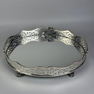 Könnte beinhalten: Ein rundes, silberfarbenes Spiegeltablett mit einem filigranen, durchbrochenen Rand und einer gewellten Kante. Das Tablett steht auf drei kleinen, verzierten Füßen und besitzt eine reflektierende Glasfläche, die die Umgebung spiegelt.