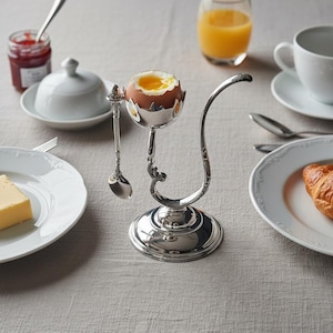 May include: A soft-boiled egg in a silver egg cup with a decorative handle and spoon. The breakfast table also includes a glass of orange juice, a cup of coffee, a plate with a croissant, a plate with butter, and a jar of jam.