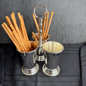 Puede incluir: Soporte de metal plateado con tres tazas para sostener palitos de pan. El soporte tiene un asa de anillo y está lleno de palitos de pan.