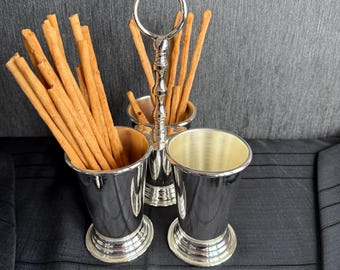 Puede incluir: Soporte de metal plateado con tres tazas para sostener palitos de pan. El soporte tiene un asa de anillo y está lleno de palitos de pan.