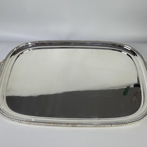 Puede incluir: Una bandeja de servicio rectangular de color plateado con esquinas redondeadas y una superficie pulida y reflectante. La bandeja presenta un borde decorativo con cuentas y dos asas curvas y ornamentadas en los lados opuestos.