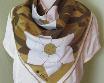 May include: A square silk scarf featuring a large white flower with a yellow center and brown and gold floral accents. The scarf has a white border with the signature "Usna" and a small insect graphic. The scarf is draped over a mannequin.
