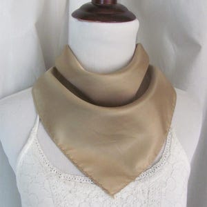 May include: A light brown silk scarf with a triangular shape. The scarf is folded and displayed on a white lace top.