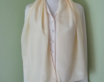 May include: A pale yellow silk scarf draped around a white blouse with buttons. The scarf is long and flowing, with a simple design.