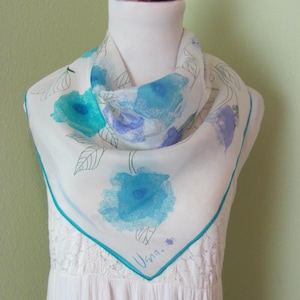Peut inclure: Un foulard en soie blanc avec un motif floral dans les tons de bleu et de turquoise. Le foulard a une bordure turquoise et la signature "Vana" dans le coin. Le foulard est drapé sur un mannequin.
