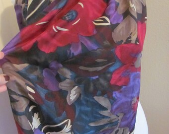 Op de afbeelding: Een sjaal met bloemenpatroon in rood, paars, blauw en bruin. De sjaal is over een witte mannequin gedrapeerd, wat de val en het patroon van de stof benadrukt. Het ontwerp suggereert een vintage of artistieke stijl.