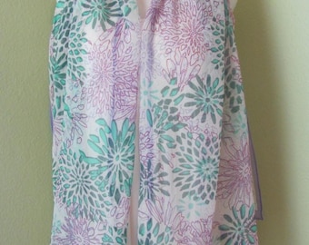 May include: A sheer scarf with a floral pattern in shades of purple and teal on a white background. The scarf is draped over a white form, showcasing its delicate fabric and design. The edges are trimmed with a lavender border.